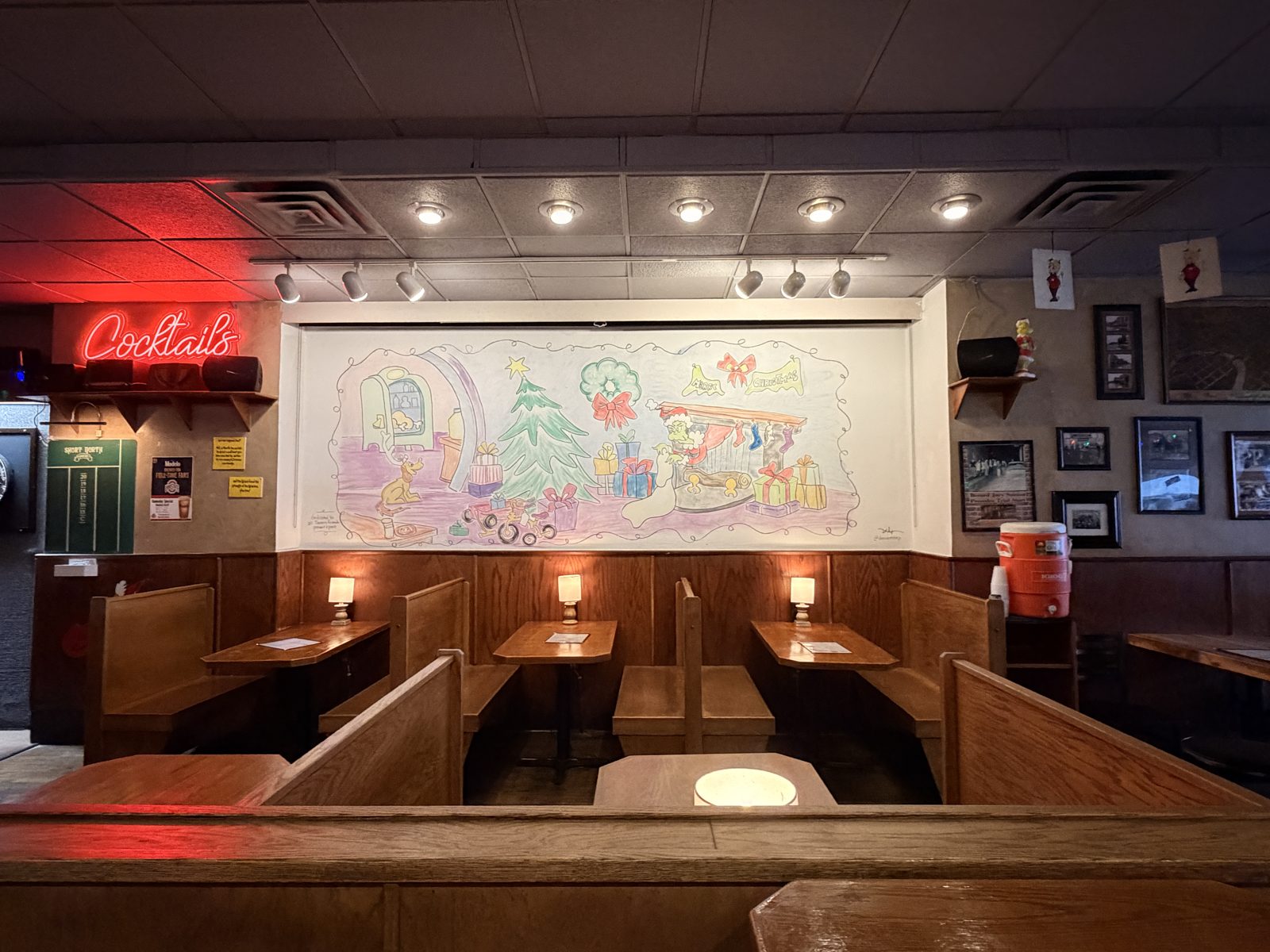 Wooden booths under a hand-painted holiday mural and a red Cocktails neon sign at a Short North bar