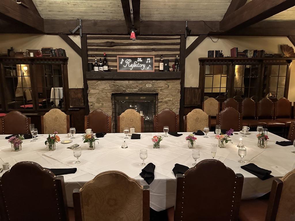 A long table set for dinner at The Refectory beneath exposed beams and a stone fireplace